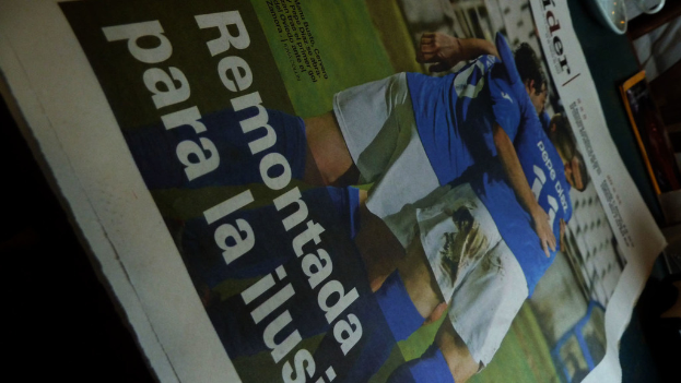 Eine Zeitung auf einem Tisch zeigt drei Männer in blauen T-Shirts, die möglicherweise Fußball spielen, mit Text am unteren Bildrand.