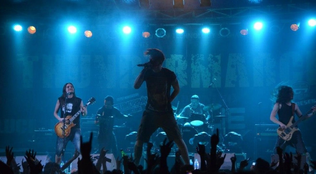 Eine Gruppe von Musikern, die auf einer hell erleuchteten Bühne spielen, einige halten Gitarren, einer hält ein Mikrofon und ein anderer spielt Schlagzeug im Hintergrund, während Zuschauer im Vordergrund die Hände heben.