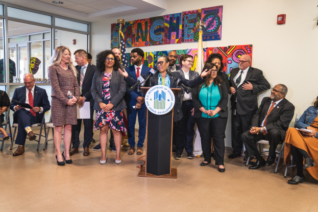 Eine Gruppe von Menschen, die vor einem Podium mit einem Mikrofon und einer Tafel mit Text stehen und sitzen, mit Flaggen, einer Tafel, einem Feuerlöscher und Deckenleuchten an der Rückwand.