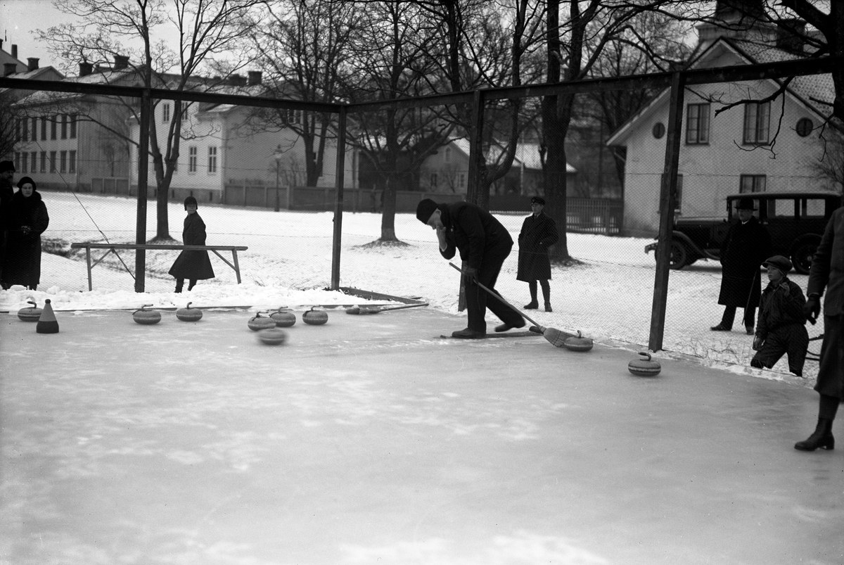 Schwarzes Bild von Menschen, die Curling auf einer Outdoor-Eisbahn spielen, umgeben von einem Zaun, einer Bank, Bäumen, Gebäuden und einem geparkten Fahrzeug im Hintergrund.