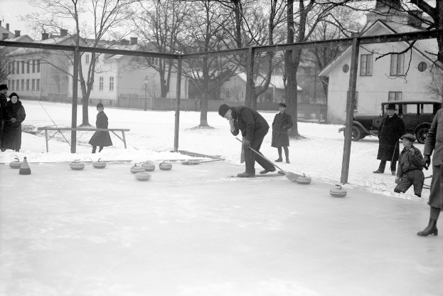 Schwarzes Bild von Menschen, die Curling auf einer Outdoor-Eisbahn spielen, umgeben von einem Zaun, einer Bank, Bäumen, Gebäuden und einem geparkten Fahrzeug im Hintergrund.
