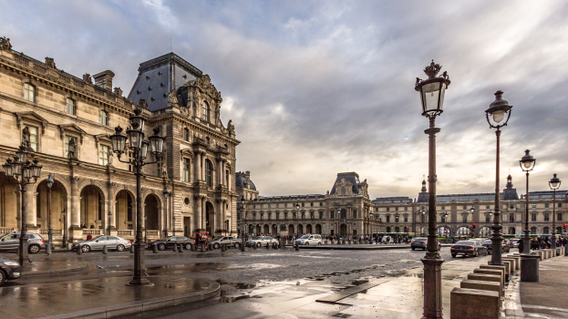 Außenansicht des Louvre-Museums in Paris, das seine charakteristische Architektur, Laternenpfähle, Lichter, fahrende Fahrzeuge, Passanten auf dem Gehweg und eine bewölkte Himmel zeigt.