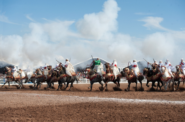 Menschen reiten auf dem Boden mit anderen, Zäune und Unterkünfte im Hintergrund unter einem bewölkten Himmel.