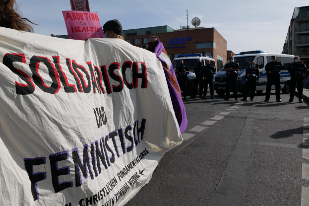 Eine Gruppe von Menschen marschiert auf der Straße, hält ein Banner mit der Aufschrift "Solidarität und Feminismus", mit parkenden Fahrzeugen, Gebäuden, einer Schüsselantenne und einem klaren blauen Himmel im Hintergrund.