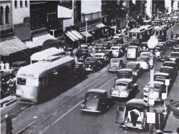 Vollständige Stadtstraße mit mehreren Autos, Bussen, Fußgängern, Gebäuden und Strommasten; unten sichtbarer Text.