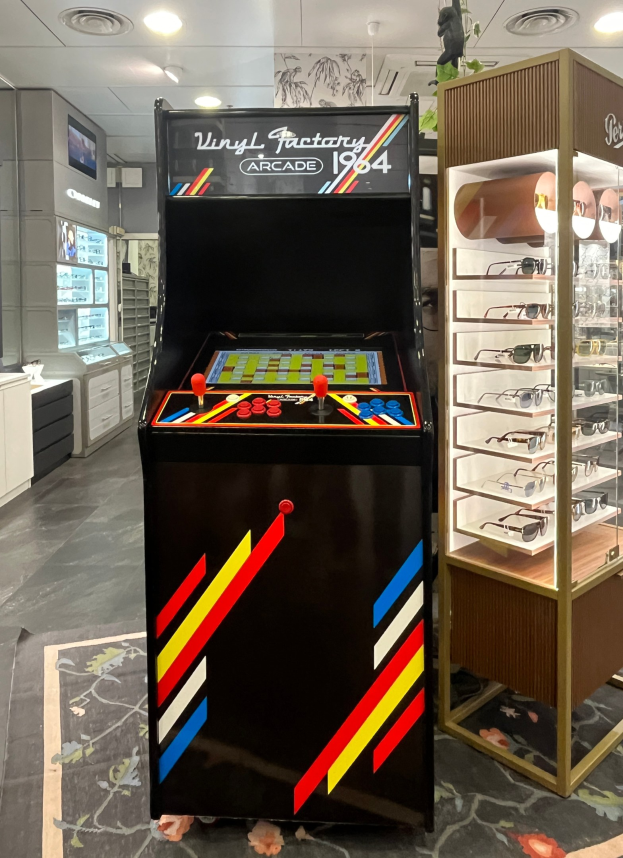 Ein Retro-Laden mit einem alten Arcade-Automaten in der Mitte, daneben ein Gestell mit Gläsern, Schränke, einen Fernseher und Deckenleuchten im Hintergrund.