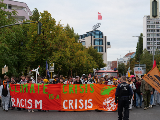 Menschen marschieren eine baumbestandene Straße entlang und halten ein 'Klima-Krise ist eine Krise'-Schild hoch mit parkenden Fahrzeugen, Gebäuden und einem klaren Himmel im Hintergrund.