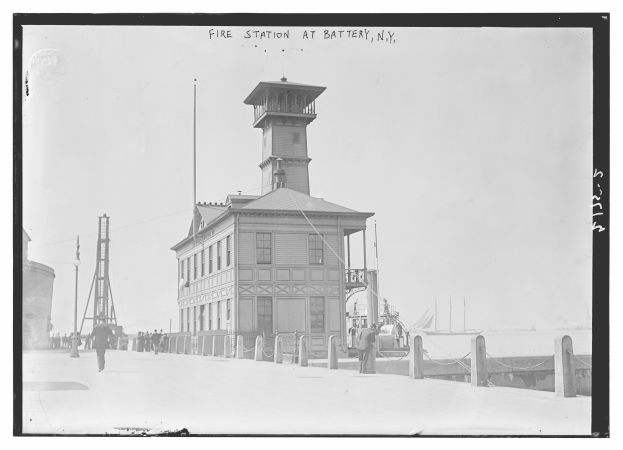 Ein Schwarz-Weiß-Foto einer Feuerwache in Battery, New York, mit einem Gebäude, Menschen, Pfählen, einem Zaun, Booten auf dem Wasser und einem bewölkten Himmel.