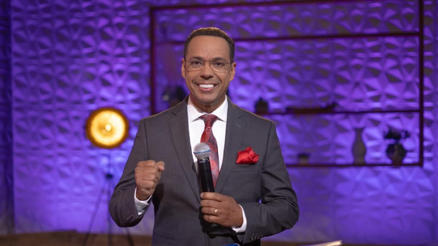 Stephen Colbert, Moderator von *The Daily Show with Trevor Noah*, steht auf einer Bühne und hält ein Mikrofon in der Hand, trägt einen Anzug, Krawatte und eine Brille, wobei ein Licht und ein Ständer hinter ihm sichtbar sind.