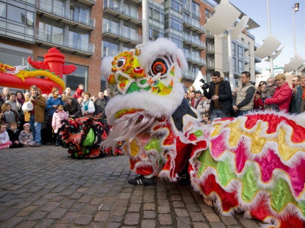 Ein farbenfrohes chinesisches Neujahrsfest in Amsterdam mit einer Löwen-Tanzvorstellung vor einer Zuschauermenge, einige halten Kameras, vor einem Hintergrund aus Gebäuden, Laternenmasten und einem klaren blauen Himmel.