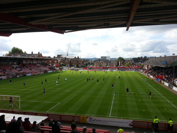 Ein Fußballspiel wird im St Mary's Stadium gespielt, mit Zuschauern auf den Bänken und Stehplätzen, einem Torpfosten auf der linken Seite und Gebäuden, Bäumen und einem bewölkten Himmel im Hintergrund.