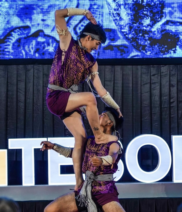 Zwei Performer in violetten Kleidern führen einen akrobatischen Hebemanöver auf der Bühne vor einem Publikum aus, mit einem schwarzen Vorhang und einem Bildschirm im Hintergrund.