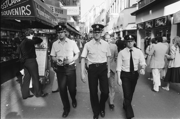 Drei Polizisten in Uniform gehen eine belebte städtische Straße entlang, die von hohen Gebäuden und Geschäften gesäumt ist, mit Passanten und einem sichtbaren Himmel im Hintergrund.