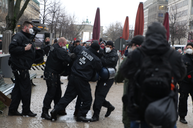 Eine Gruppe von Polizeibeamten in schwarzer Einsatzkleidung und Masken, die eine Straße in Berlin, Deutschland, entlanggehen, mit Gebäuden, Bäumen, Schirmen, Masten, Fahrzeugen und einem sichtbaren Himmel im Hintergrund.