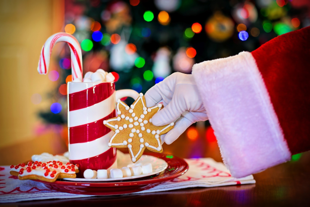 Mann in Santa Claus Mütze mit einem Becher heißer Schokolade und einem Zuckerstange haltend, mit einem Teller mit Keksen und einem Tuch auf dem Tisch, vor einem Weihnachtsbaum und einer Wand.