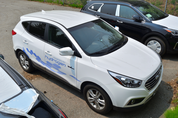 Drei Autos nebeneinander auf einem Parkplatz geparkt, mit einem Maschendrahtzaun und Gras im Hintergrund, auf einem Auto steht der Text 'wasserstoffbetriebenes Fahrzeug'.