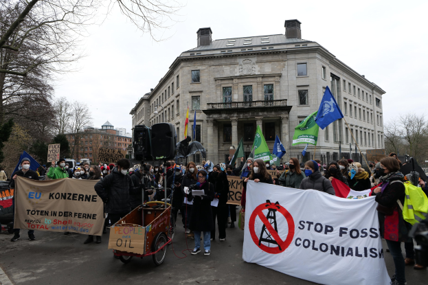 Eine große Gruppe von Menschen marschiert bei einer Demonstration gegen fossile Brennstoffe, trägt Schilder und Fahnen, mit einem Fahrzeug im Vordergrund und Gebäuden, Bäumen und einem klaren blauen Himmel im Hintergrund.