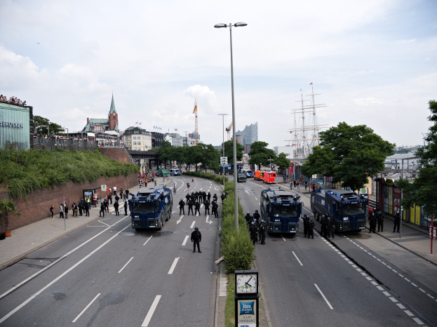 Eine große Gruppe von Polizeibeamten steht auf der Seite einer Straße, umgeben von Fahrzeugen und Menschen, mit Laternen, Schildern mit Text, Pflanzen, Bäumen, Gebäuden und Türmen im Hintergrund unter einem bewölkten Himmel.