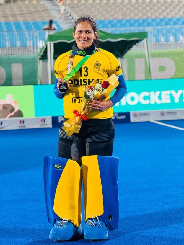 Frau in gelb-schwarzem Kleid mit Blumenstrauß und Medaille auf blauem Feld, mit Zuschauern im Hintergrund, die einen Hockey-Sieg feiern.