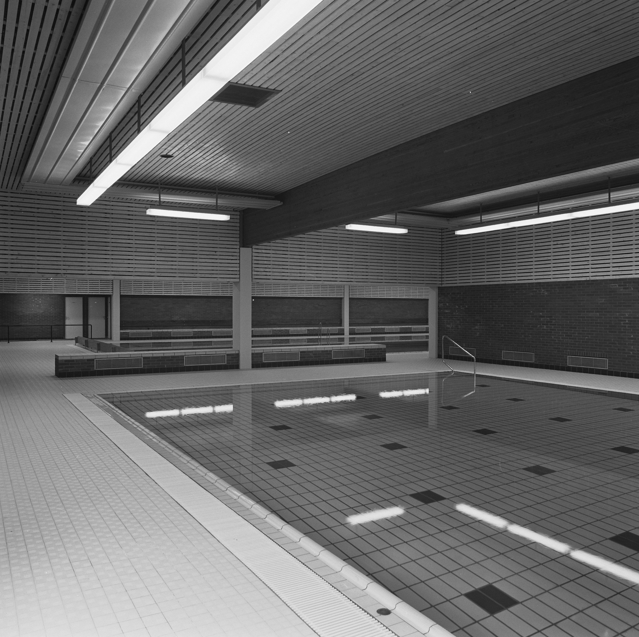 Indoor-Schwimmbad mit klarem Wasser, gefliesten Wänden und Boden, Deckenbeleuchtung und minimalen Umgebungsobjekten in einem großen Gebäude.