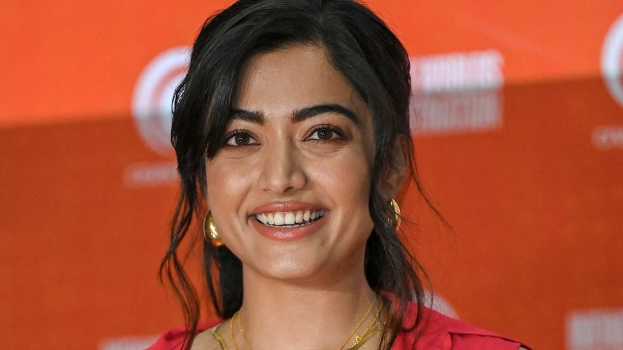 Eine Frau in einem leuchtend roten Kleid mit einem eleganten Dutt und dezentem Make-up posiert strahlend vor der Kamera, trägt schwarze High Heels und hat ein hellgelbes Banner mit der Aufschrift "Sonam Kapoors Schönste Looks vom Cannes Film Festival" im Hintergrund.