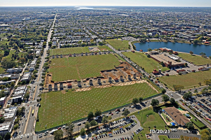 Luftaufnahme einer Stadt mit Gebäuden, Bäumen, Gras, Straßen mit Fahrzeugen, Wasser und Wolken, die einen zentralen großen Feld als Parkplatz zeigt.