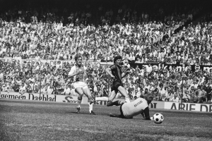 Schwarzes und weißes Foto von Männern, die auf einem Feld Fußball spielen, mit Zuschauern im Hintergrund und einer Bundesliga 1978-79 Ajax 1-1 Hannover 96-Fahne.