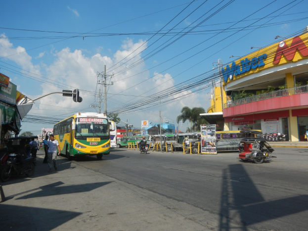 Eine belebte Stadtstraße mit Fahrzeugen wie einem Bus und Motorrädern, Fußgängern auf den Gehwegen, Verkehrsampeln, Strommasten, Gebäuden mit Namensschildern, Bäumen und einem bewölkten Himmel.
