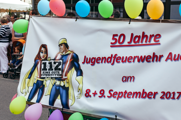 Eine Gruppe von Menschen auf der Straße mit einem Banner, auf dem "50 Jahre Jugendfeuerwehr August 2017" steht, mit Kinderwagen, Ballons, einem Zelt, einem Gebäude und Bäumen im Hintergrund.