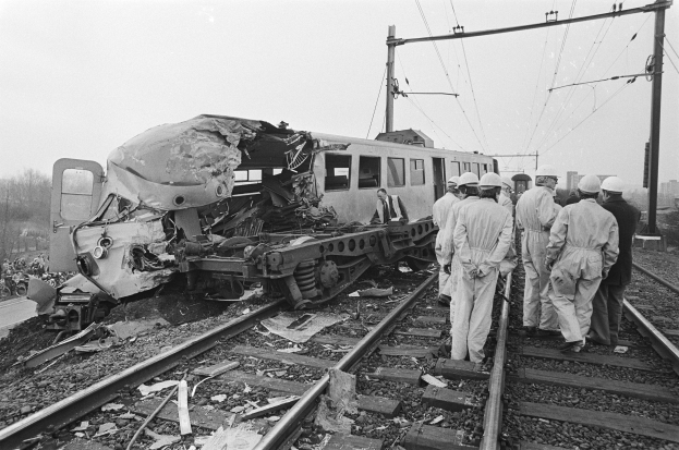 Ein Schwarz-Weiß-Bild von einer Zugkollision auf den Schienen mit einer Gruppe von Helmpersonen in der Nähe, vor einem Hintergrund von Strommasten, Drähten, Bäumen, Gebäuden und einem klaren Himmel.