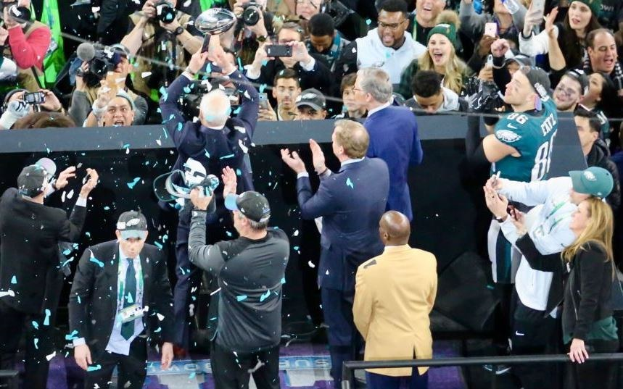 Philadelphia Eagles Spieler feiern mit dem Vince Lombardi Trophy, tragen Kappen, halten den Pokal und werfen Konfetti, mit einem Geländer und kameraschwingenden Personen im Hintergrund.