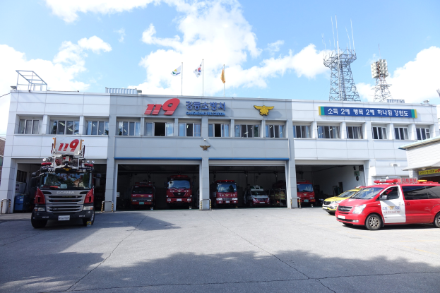 Feuerwache mit mehreren Feuerwehrautos vor dem Gebäude geparkt, Gebäude mit Fenstern und Text, Flaggen mit Stangen im Hintergrund, bewölkter Himmel und eine ferne Turm.