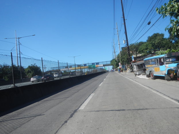 Straßenverkehr mit fahrenden Autos, geparktem Bus, Strommasten mit Drähten, einem Zaun auf der linken Seite, Bäumen, Straßenlaternen, einer Brücke im Hintergrund und einem blauen Himmel.