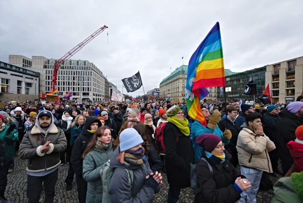 Eine große Gruppe von Menschen bei einer LGBTQ+-Rechtsdemo in Berlin, die Fahnen und Plakate schwenken, mit Gebäuden, einem Kran und Wolken im Hintergrund.