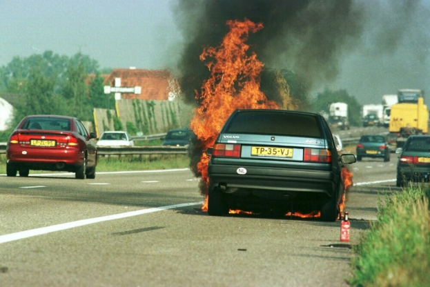 Ein Auto, das in Flammen steht, am Straßenrand mit anderen Fahrzeugen in der Nähe, Bäumen, Gebäuden und einem klaren blauen Himmel im Hintergrund und einem Feuerlöscher auf der rechten Seite des Bildes.