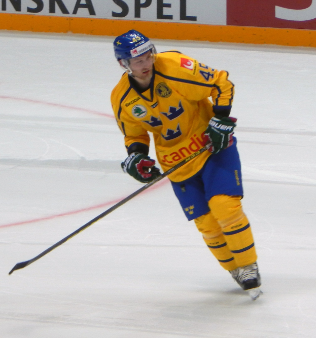 Ein Hockey-Spieler in Helm, Handschuhen und Schlittschuhen hält einen Hockey-Schläger auf dem Eis, mit einer Tafel im Hintergrund, auf der Text steht.