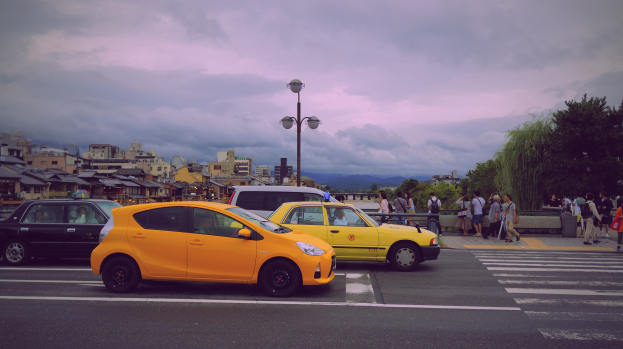 Zwei gelbe Taxis stehen auf der Seite einer Stadtstraße mit Passanten, Laternen, Bäumen, Gebäuden und einem bewölkten Himmel im Hintergrund.