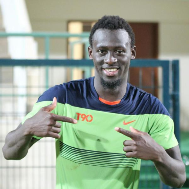 Ein Mann in einem grünen und blauen Hemd, der als Emmanuel Adebayor identifiziert wird, zeigt mit dem Finger auf die Kamera und lächelt, mit einem unscharfen Geländer, einer Wand und einer Tür im Hintergrund.