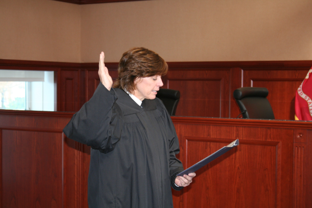 Eine Frau in einer schwarzen Robe steht vor einem Richtertisch, hält eine Akte, mit einem Mikrofon und einer Flagge auf dem Tisch und Stühlen und einem Fenster dahinter.