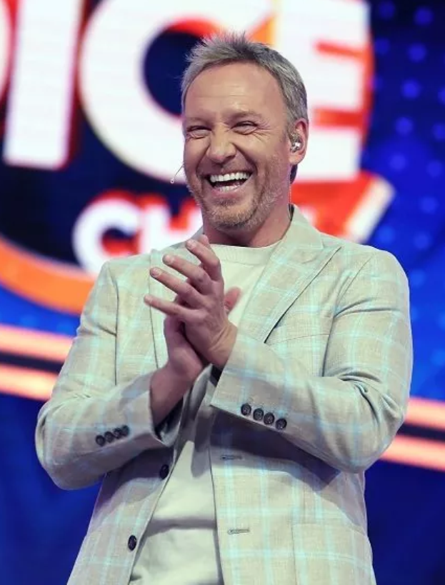 Ein Mann im Anzug steht auf einer Bühne, lächelt und klatscht, mit einem Schild im Hintergrund, auf dem 'The Voice' steht.