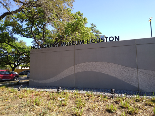 Außenansicht des Houston Holocaust Museums mit einer Gedenkwand, Steinen, Vegetation, Fahrzeugen, einem Straßenpfahl und einem bewölkten Himmel.