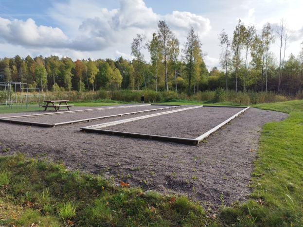 Eine grüne Wiese mit einem Picknicktisch in der Mitte, umgeben von Bäumen und einem klaren blauen Himmel, mit Spielgeräten wie Pfosten und einer Bank auf der linken Seite.
