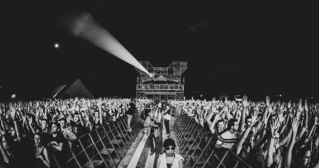 Eine Menschenmenge auf einem Konzert mit erhobenen Händen, die vor einem Geländer stehen, mit einer beleuchteten Bühne im Hintergrund, dargestellt in Schwarz-Weiß.