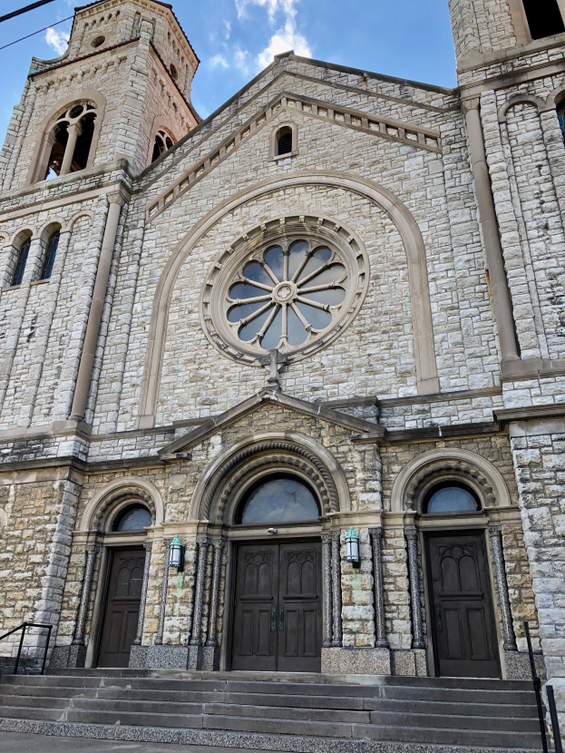 Eine große steinerne Kirche mit einem Turm, einem Glockenturm, Türen, Fenstern, Treppen, Geländern, Lampen und Drähten, die vor einem bewölkten Himmel steht.