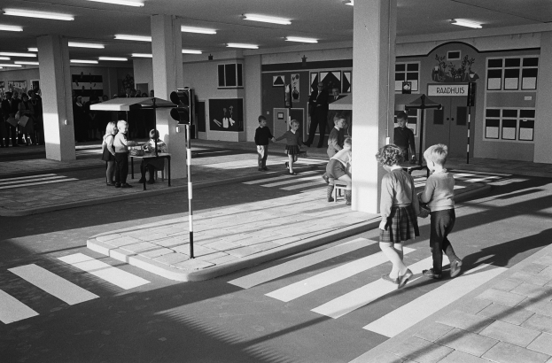 Ein Schwarz-Weiß-Foto von Kindern, die eine Straße an einer Ampel überqueren, mit Menschen, die stehen und auf Bänken sitzen, einem Verkehrssignal im Vordergrund und Säulen und Lampen im Hintergrund.