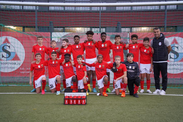 Eine Gruppe junger Männer in roten T-Shirts und schwarzen Shorts steht auf einem Fussballfeld neben einer Kiste roter Gegenstände, mit Bannern im Hintergrund, die 'FC Rot-Schwarz Eglaute - Champions League' lesen.