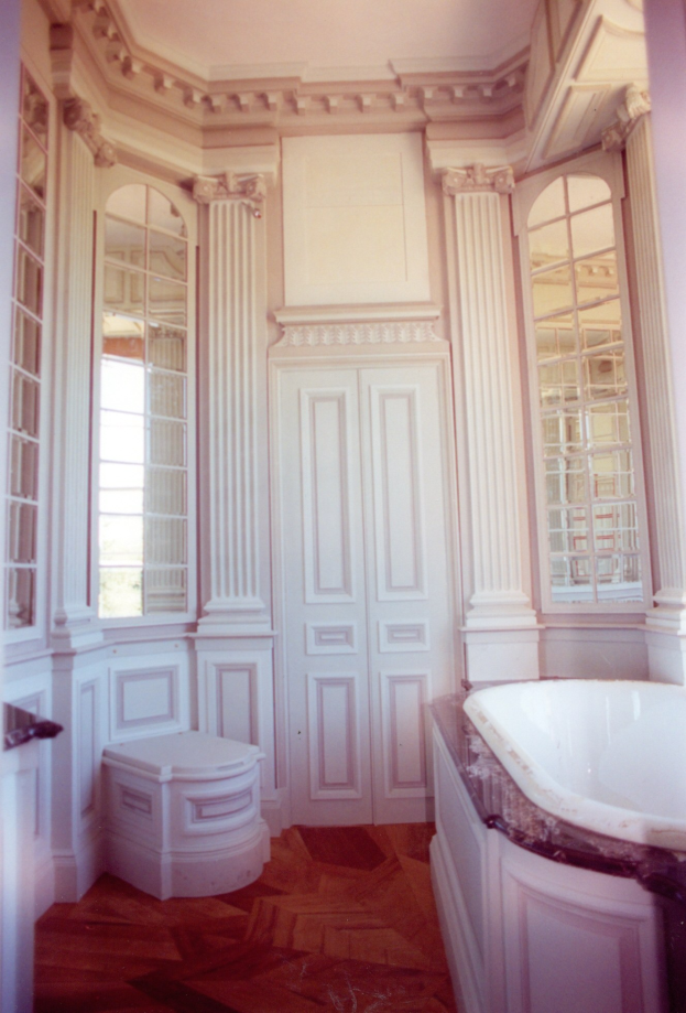 Ein luxuriöses Badezimmer mit einer großen Wanne auf der rechten Seite, einer Toilette auf der linken Seite und kunstvoll verzierten Wänden mit Fenstern und Türen im Hintergrund.