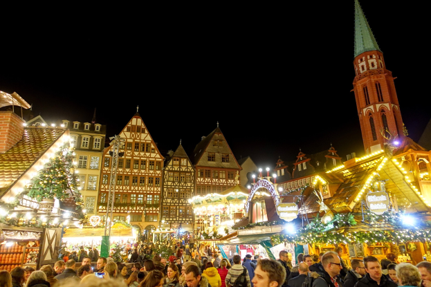 Ein geschäftiger Weihnachtsmarkt in Nürnberg, Deutschland mit Menschen um geschmückte Stände, Gebäuden mit Fenstern, einem Kirchturm und einem dunklen Himmel.
