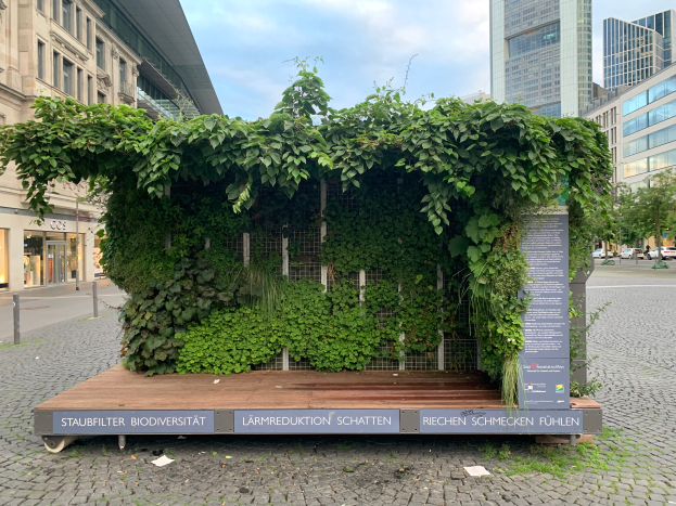 Ein grünes Gebäude mit Efeu an seiner Seite, umgeben von Bäumen, Fahrzeugen, Pfählen, Schildern und anderen Gebäuden, unter einem bewölkten Himmel, Teil der Staufbilter Biodiversität in Berlin, Deutschland.