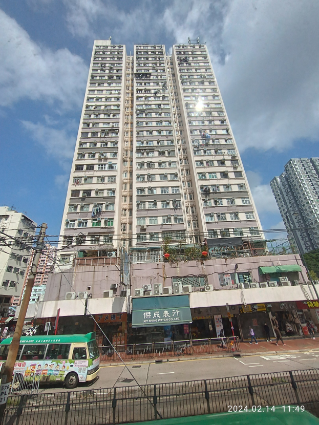 Ein hohes Wohngebäude in einer belebten Stadtstraße umgeben von anderen Gebäuden, Fahrzeugen, Fußgängern und städtischer Infrastruktur unter einem bewölkten Himmel.